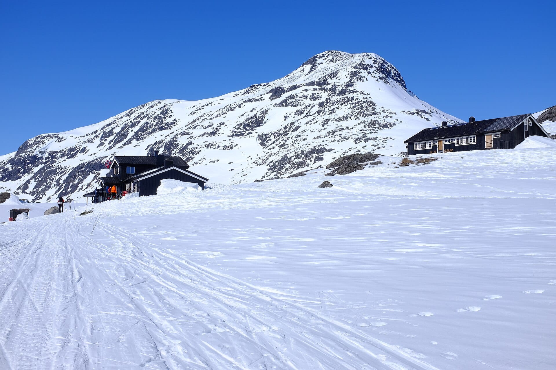 Olavsbu's dark timber cabin buildings on snow in Rauddalen, Jotunheimen, with skiers gathered at the door below a snow-covered peak