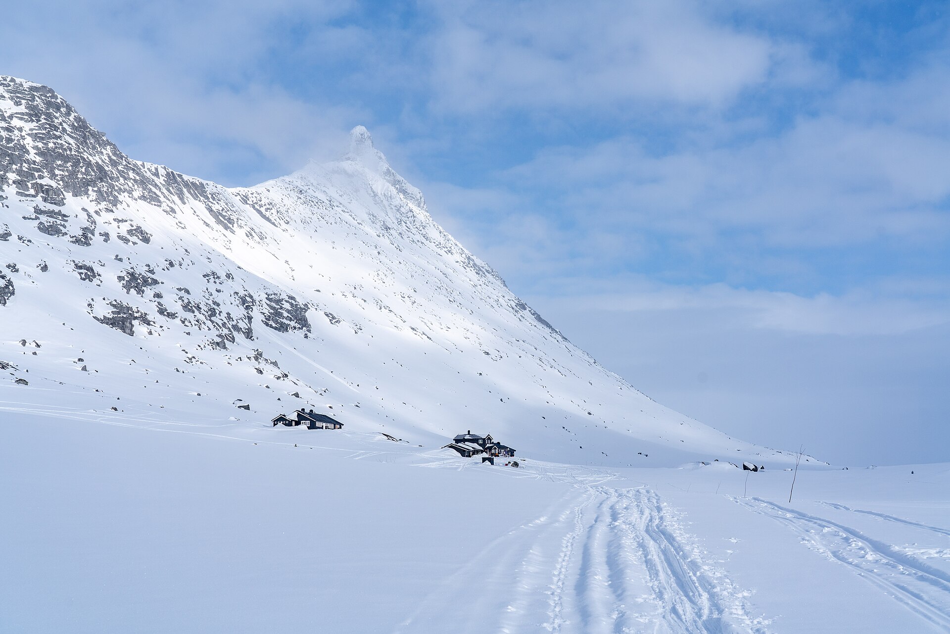 Ski tracks cross a snow-covered valley toward Olavsbu, small beneath a steep snow-covered peak in Jotunheimen