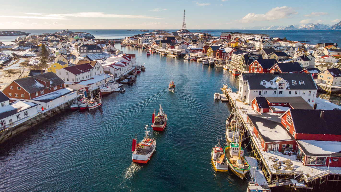 Henningsvær harbour in winter, Lofoten, with fishing boats leaving the channel between snow-covered quays and red and white wooden buildings