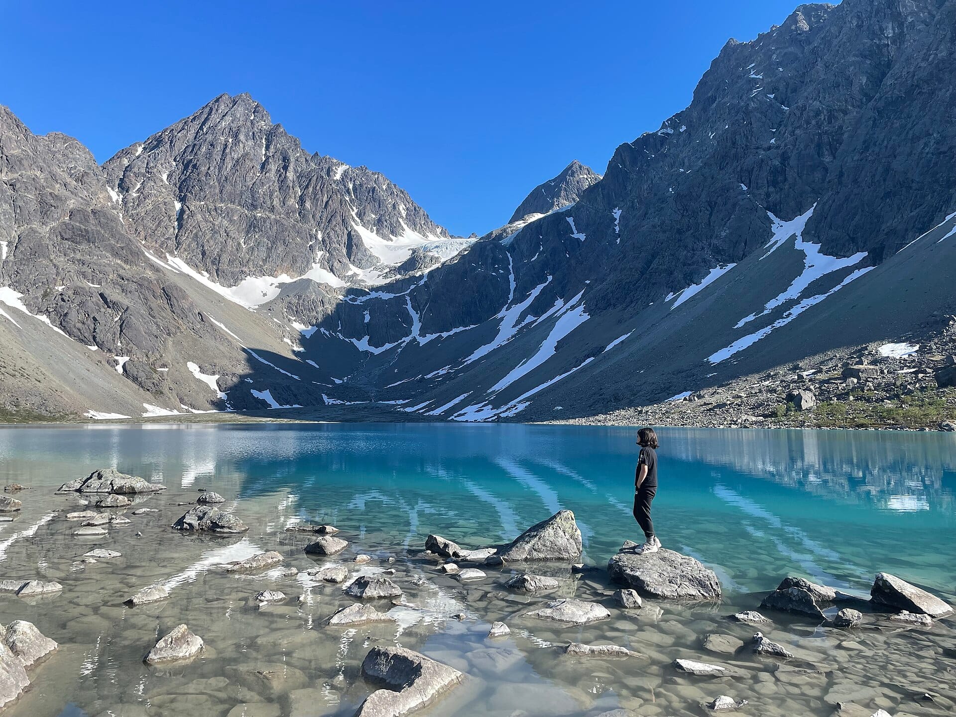 A hiker stands on a boulder at the edge of Blåisvatnet, with Lenangstinden and snow-streaked cliffs behind the lake