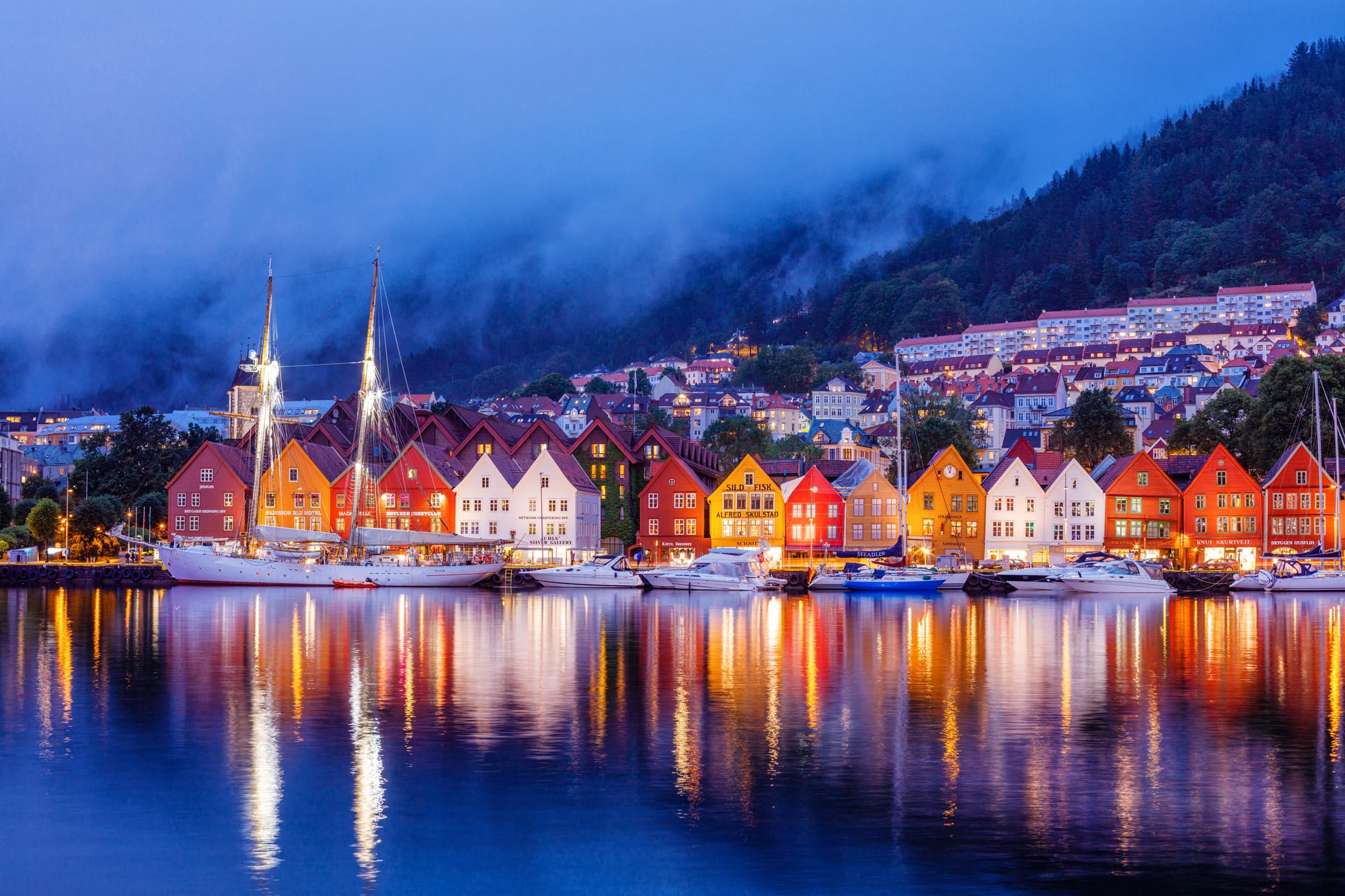 Bryggen's wooden wharf buildings lit at dusk and reflected in Bergen harbour, with cloud on the hillside behind